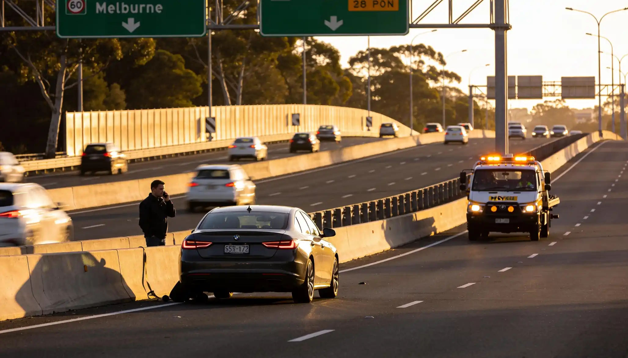 Two towing operators securing a heavily front-end damaged black car onto a flatbed tow truck on a Melbourne suburban road at golden hour sunset with residential buildings and eucalyptus trees in the background.