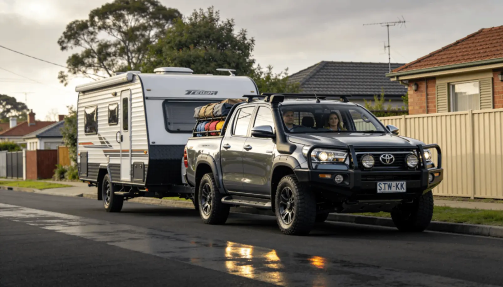 Black Toyota 4WD towing caravan on suburban street with camping gear on roof.